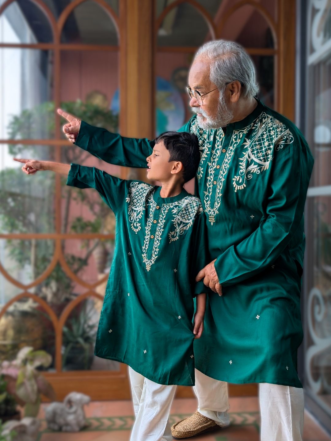 grandfather and grandson in matching traditional green embroidered kurtas standing indoors with decorative elements in the background.