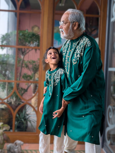 Man and child in matching emerald green traditional outfits standing indoors with decorative background