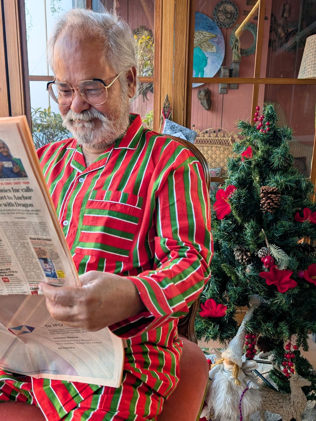 Man in red and green striped pajamas reading a newspaper next to a decorated Christmas tree.