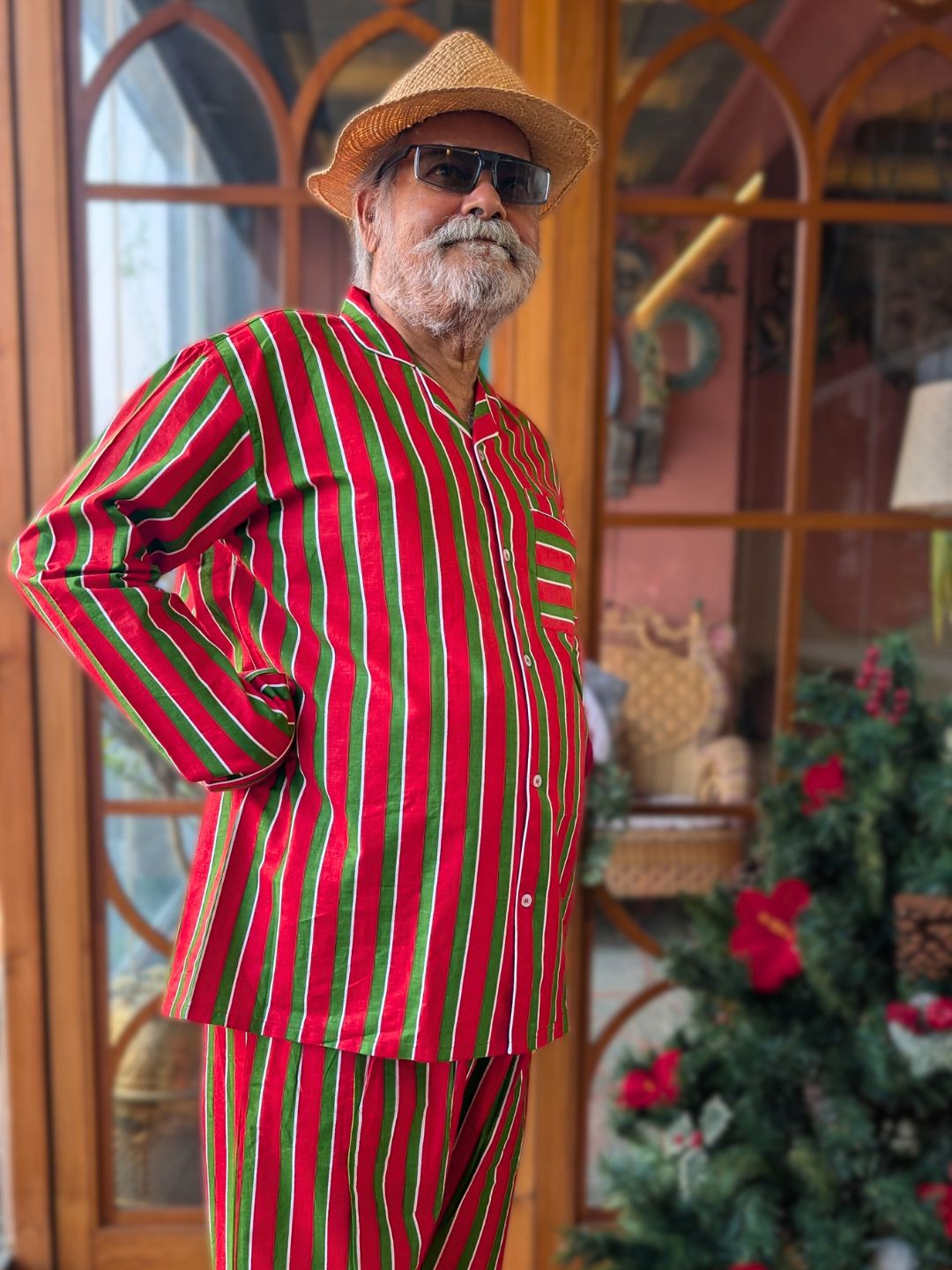 Man wearing a red and green pinstriped night suit pajama set with a straw hat in front of a decorated Christmas tree.