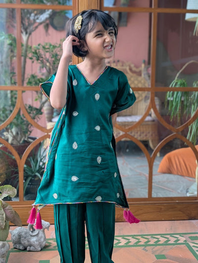 Child wearing a green traditional outfit with white patterns and pink tassels, standing indoors.