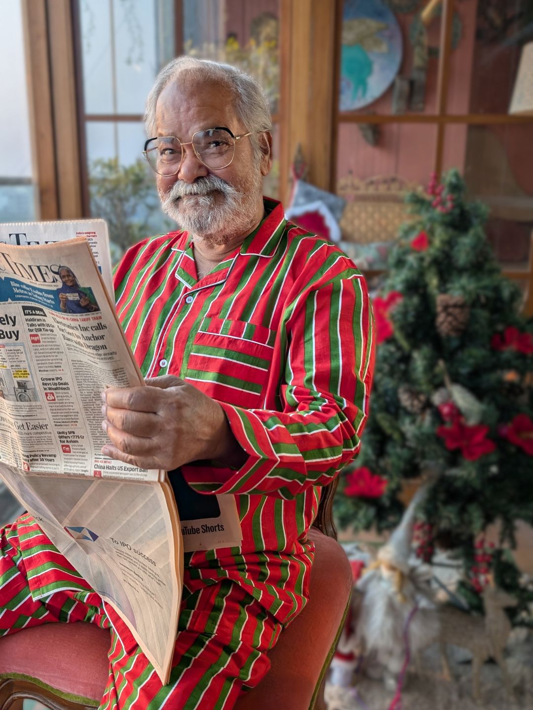 Man in red and green pajamas reading a newspaper in a festive room with a Christmas tree.