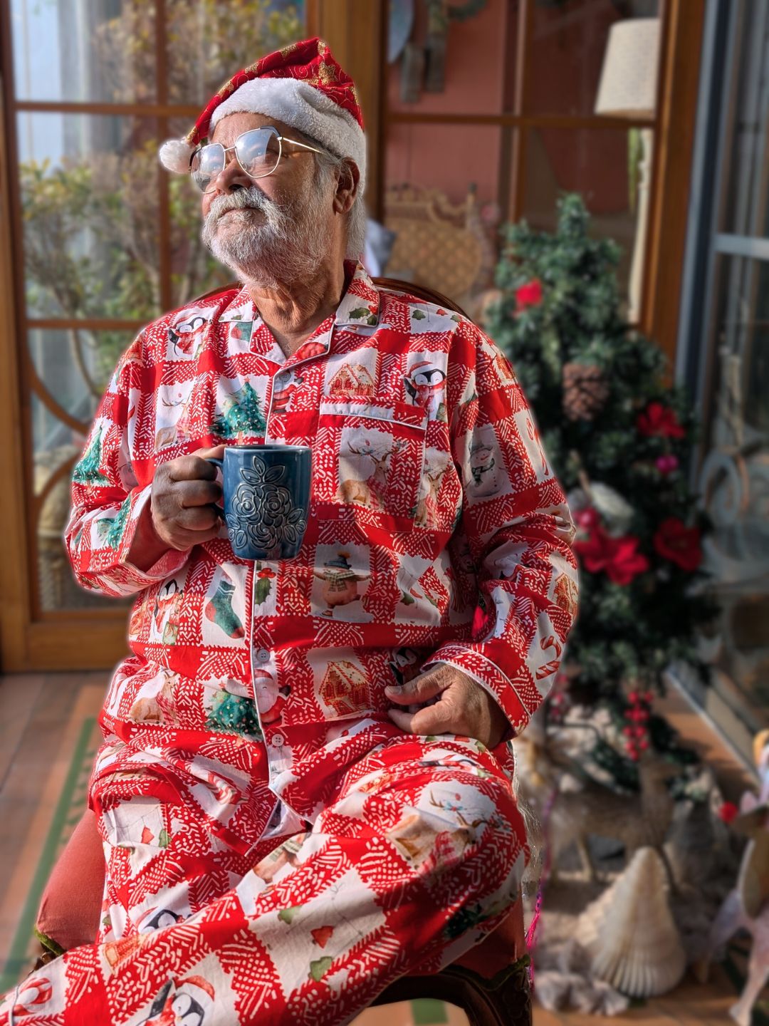 Man in Christmas night suit pajamas and Santa hat holding a mug in a cozy room with Christmas decorations.