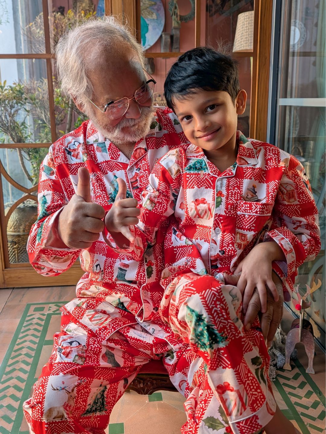 Grandpa and grandson wearing red Christmas pajamas giving thumbs up indoors.