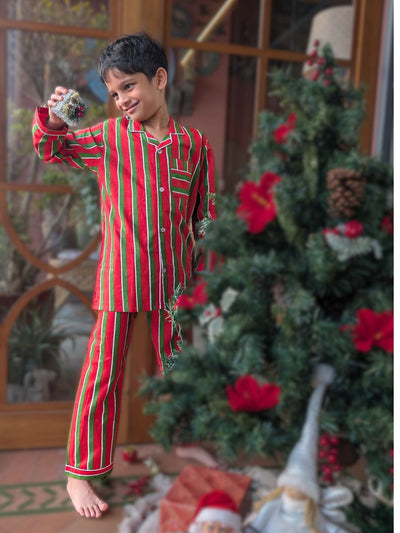 Child in red and green striped pajamas holding a Christmas ornament, standing in front of a decorated Christmas tree.