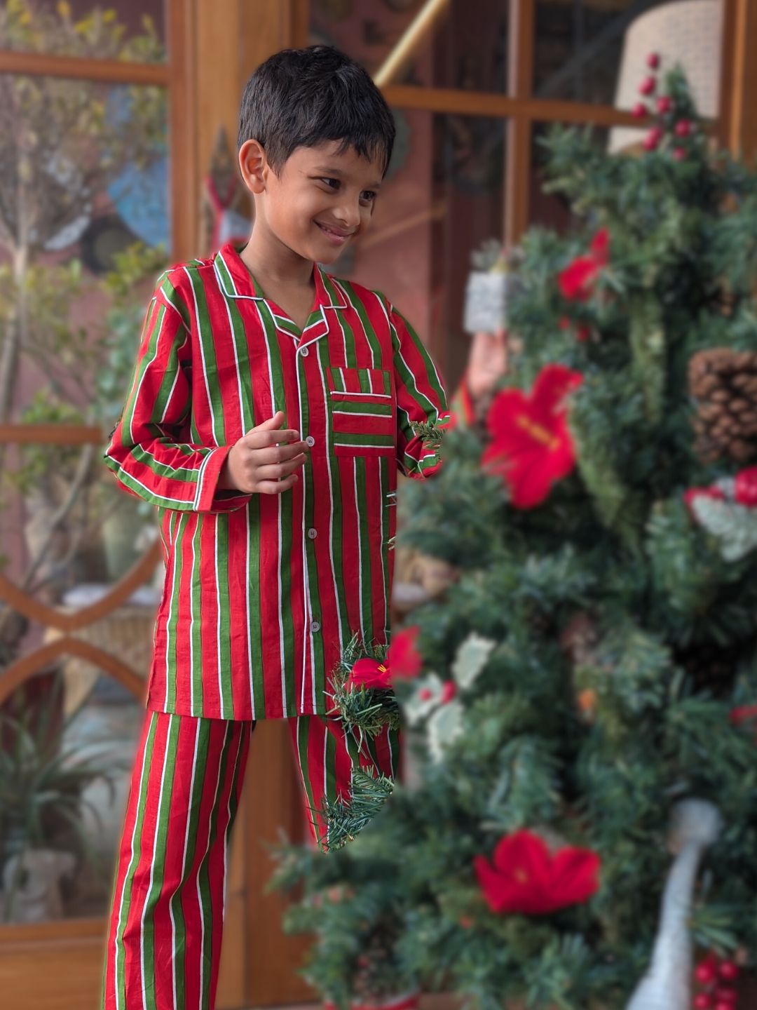Child wearing red and green striped pajamas standing next to a decorated Christmas tree.