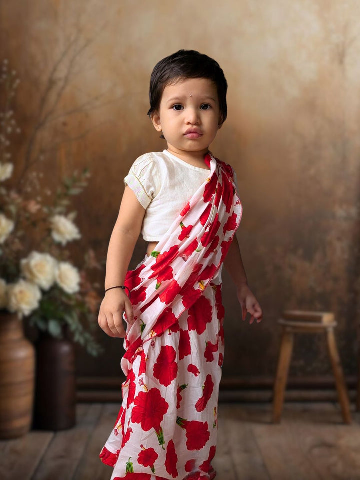 Child wearing a white saree with red floral patterns against a brown background