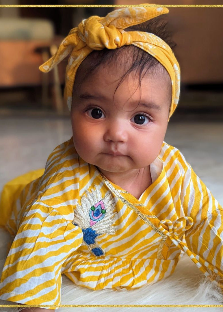 A baby girl wearing a yellow and white striped janamashtami set with embroidered krishna motif and a hairband.