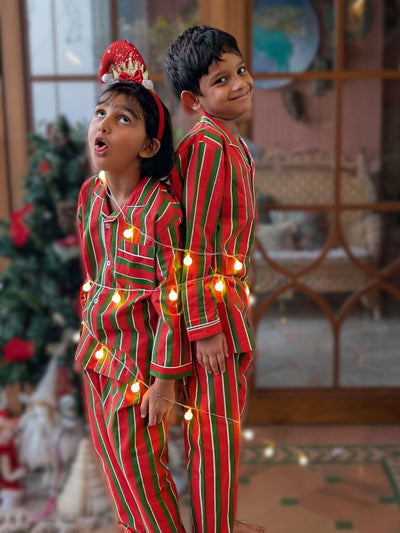 Two children in red and green striped pajamas with string lights, standing in a festive room.