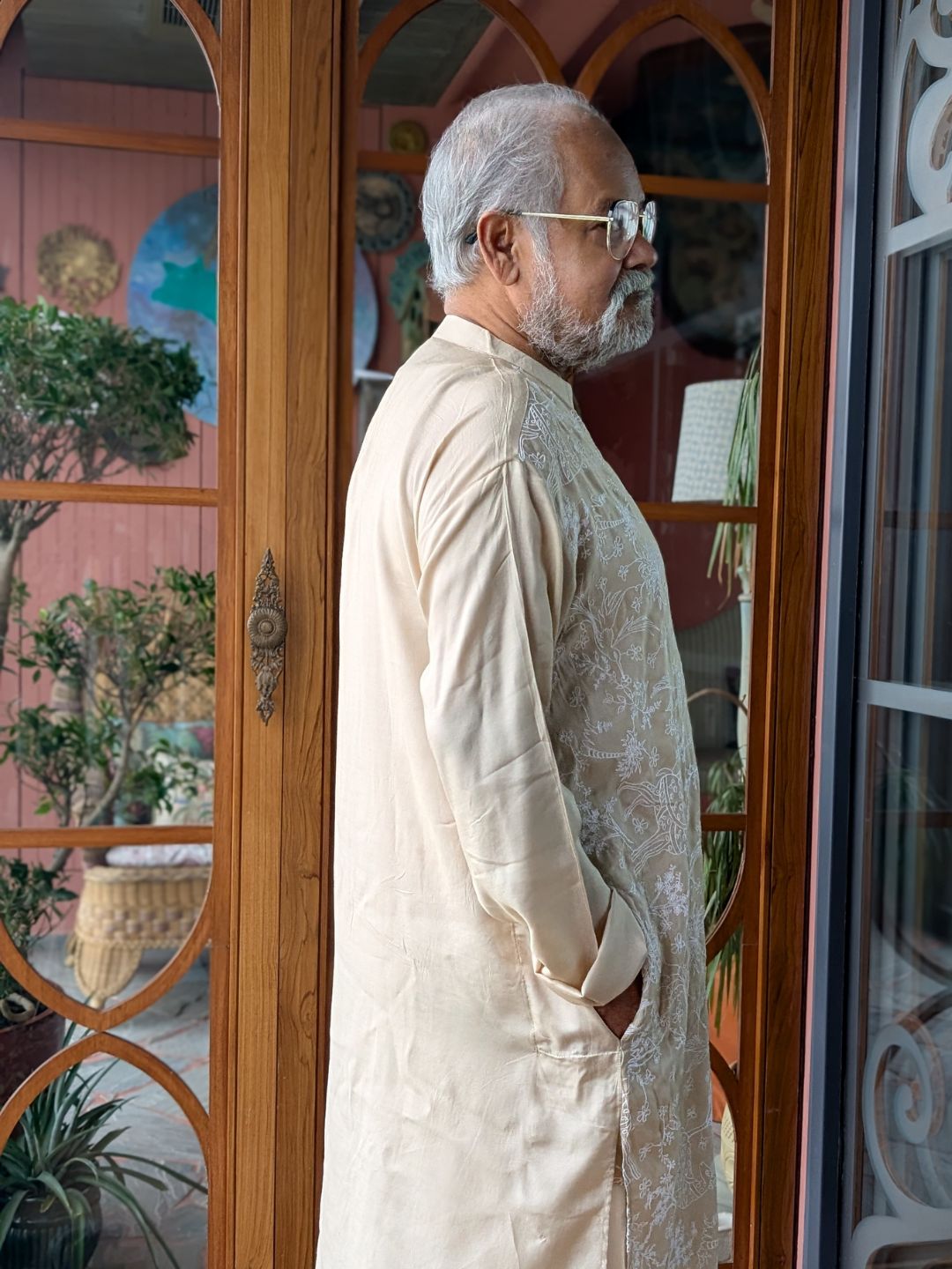 Man in a cream kurta standing in front of a decorative glass door.