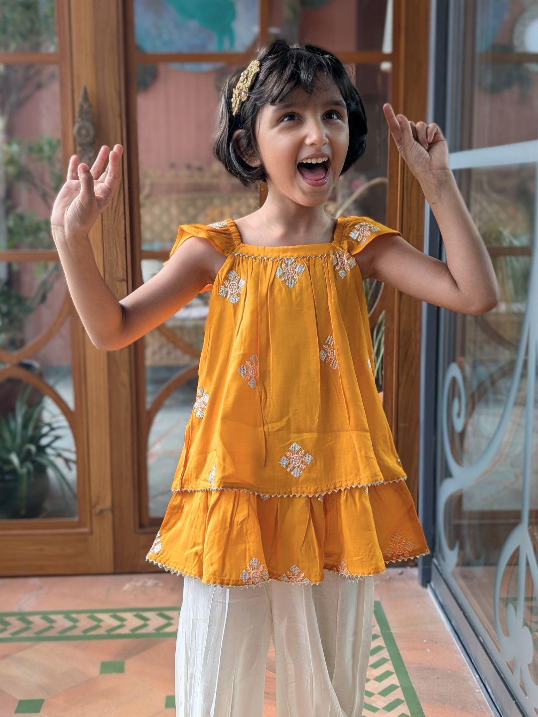 Young girl in a yellow embroidered ethnic top and white afghani pants standing indoors with a decorative background.