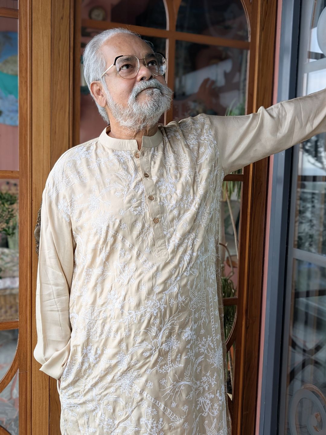 Man in a sandalwood kora coloured silk kurta standing indoors with decorative glass doors in the background