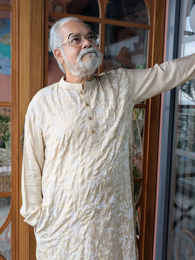 Man in a sandalwood kora coloured silk kurta standing indoors with decorative glass doors in the background