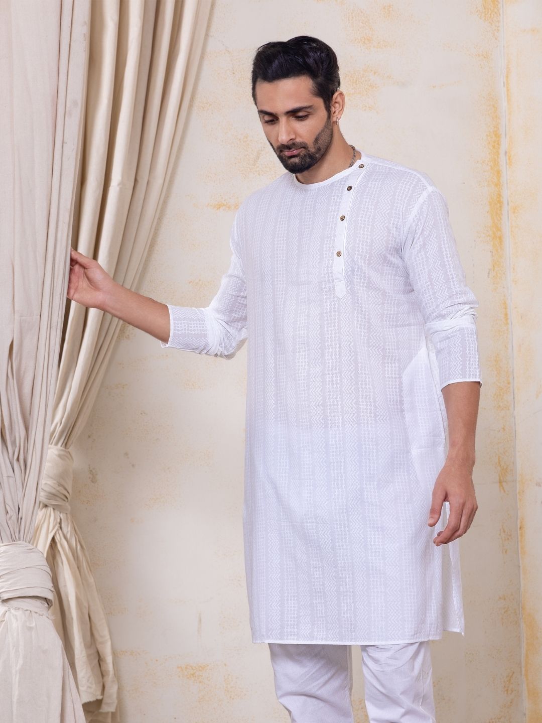 Man wearing a white kurta standing next to a beige curtain.