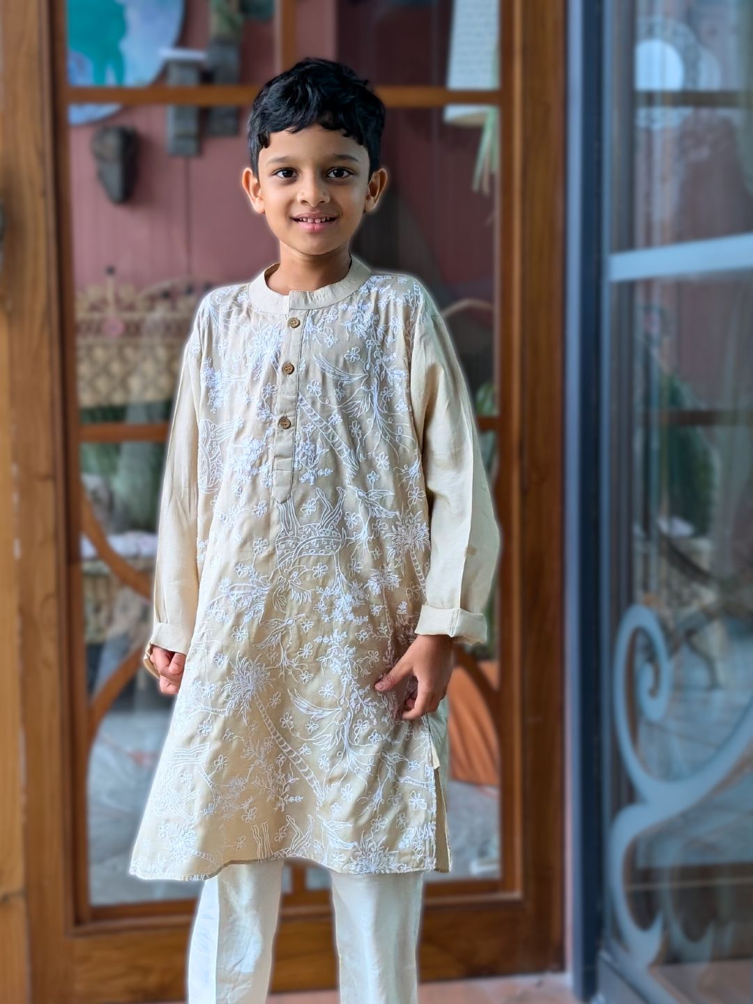 Young boy wearing a traditional cream embroidered kurta with white pajama standing in front of a glass door.