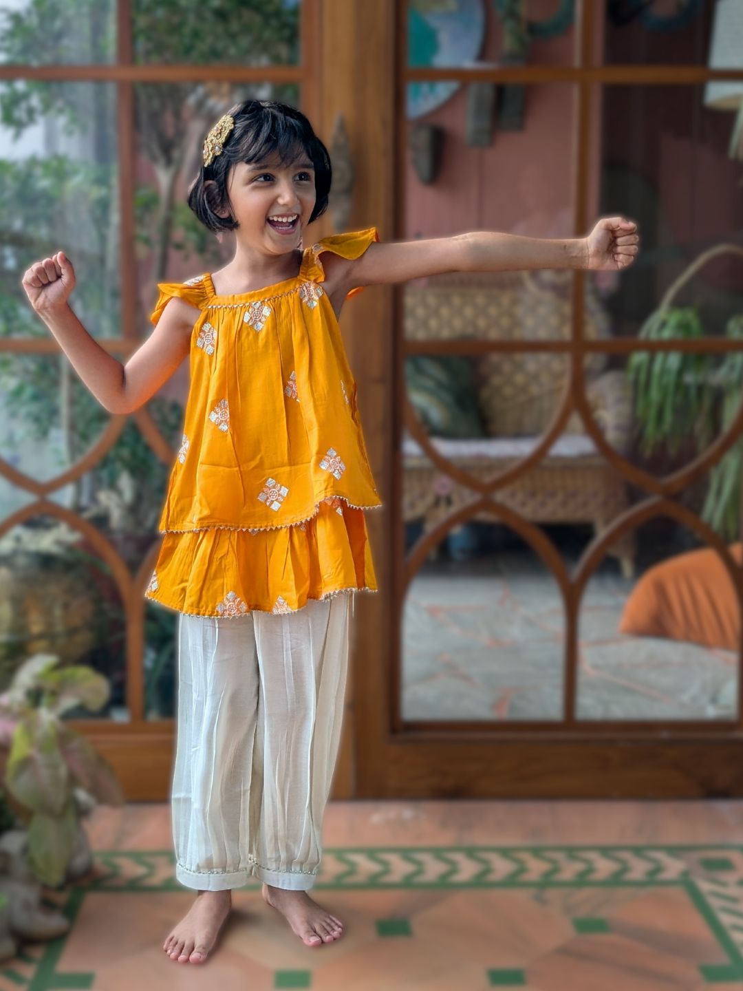 Young girl in a kesar or saffron yellow ethnic kurta top and white afghani pants standing in a room with decorative elements.