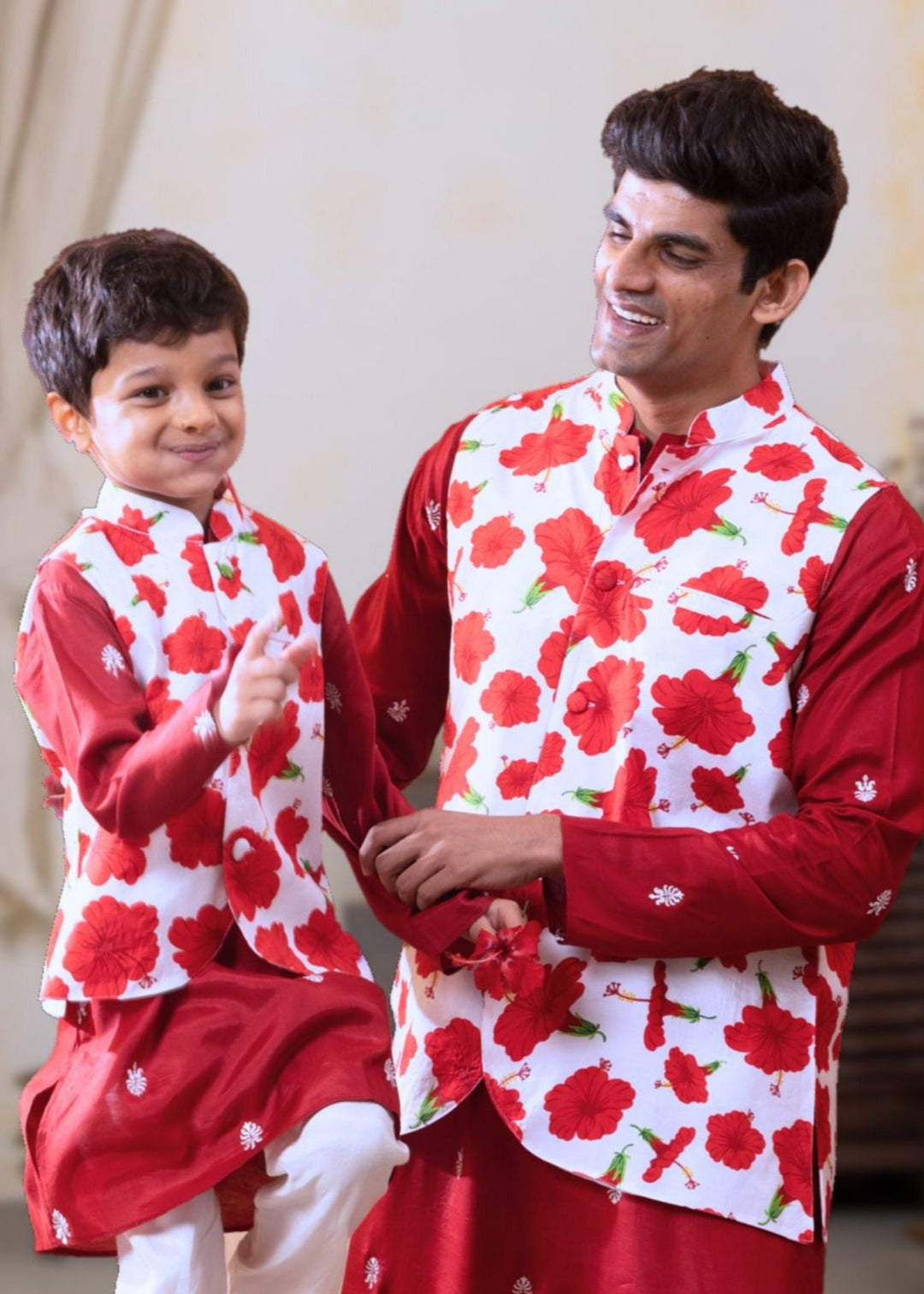Man and boy wearing matching red and white hibiscus festive floral outfits in a decorated room.