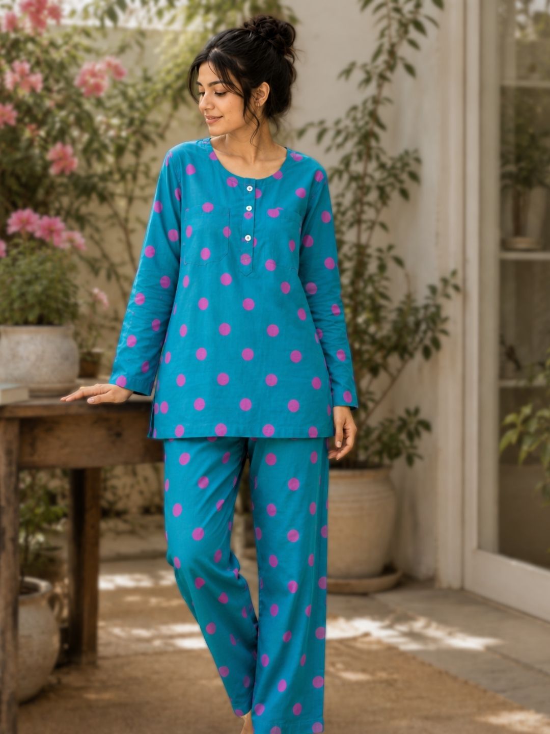 A woman wearing a teal blue two-piece no bra or braless nightsuit pajama set with pink polka dots, standing in a sunlit patio setting with potted plants and a wooden table.
