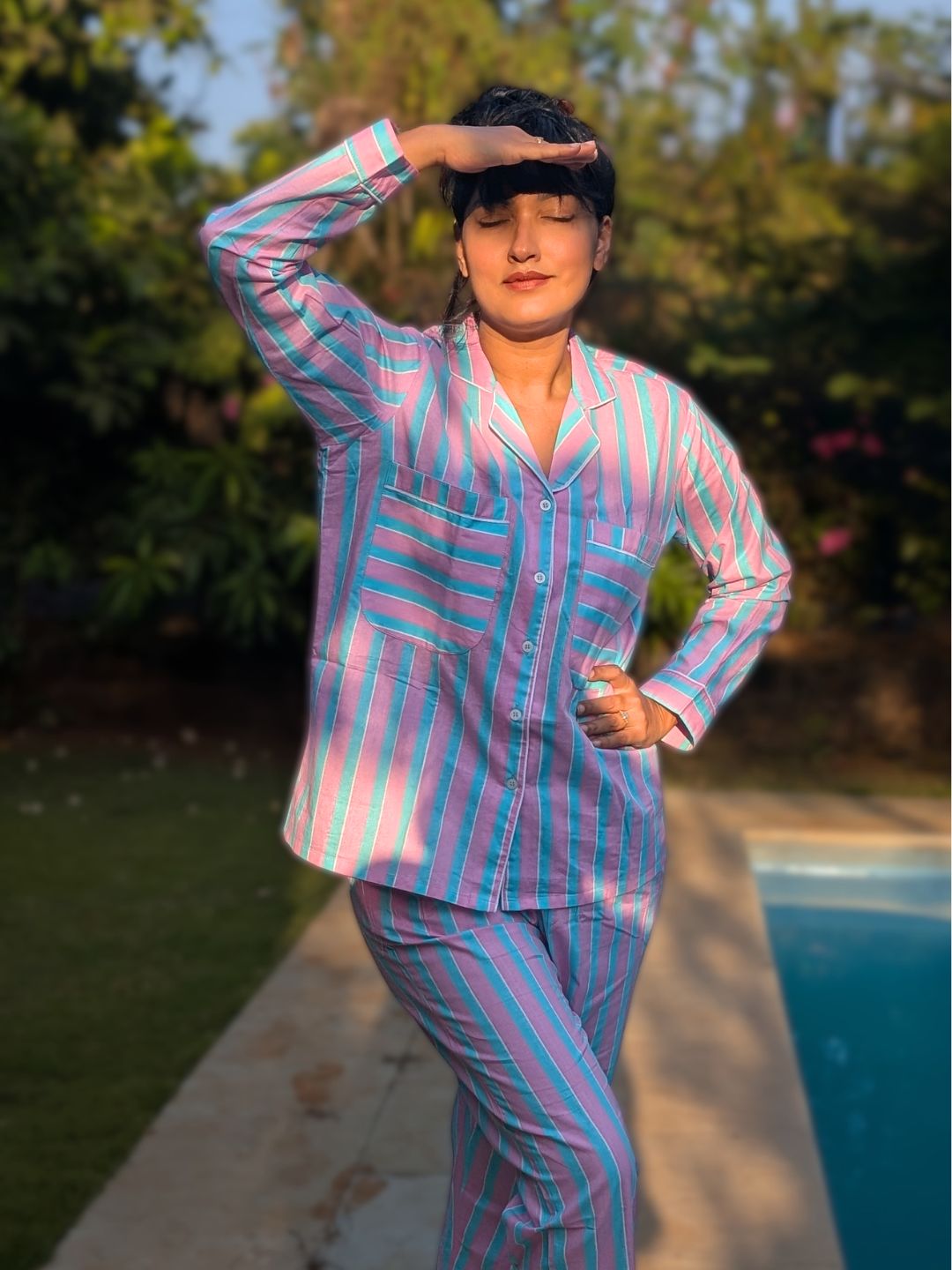woman wearing a pink and blue pinstripe cotton pajama set standing outdoors by a pool.