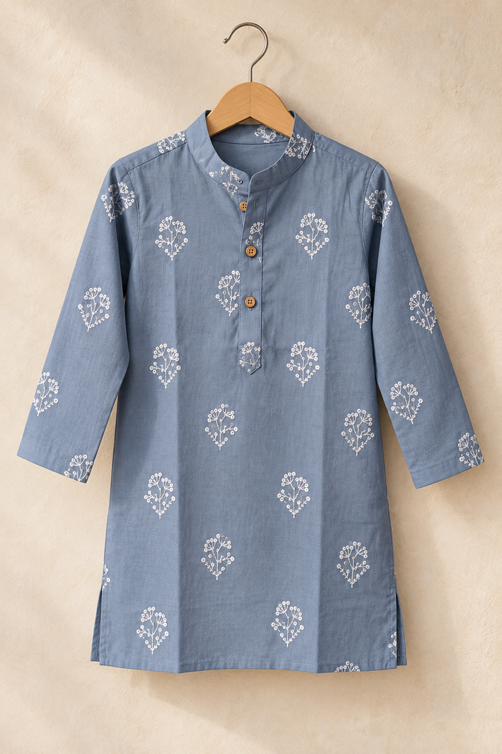 A dusty blue cotton kurta with white embroidered floral motifs, hanging on a wooden hanger against a textured beige wall.