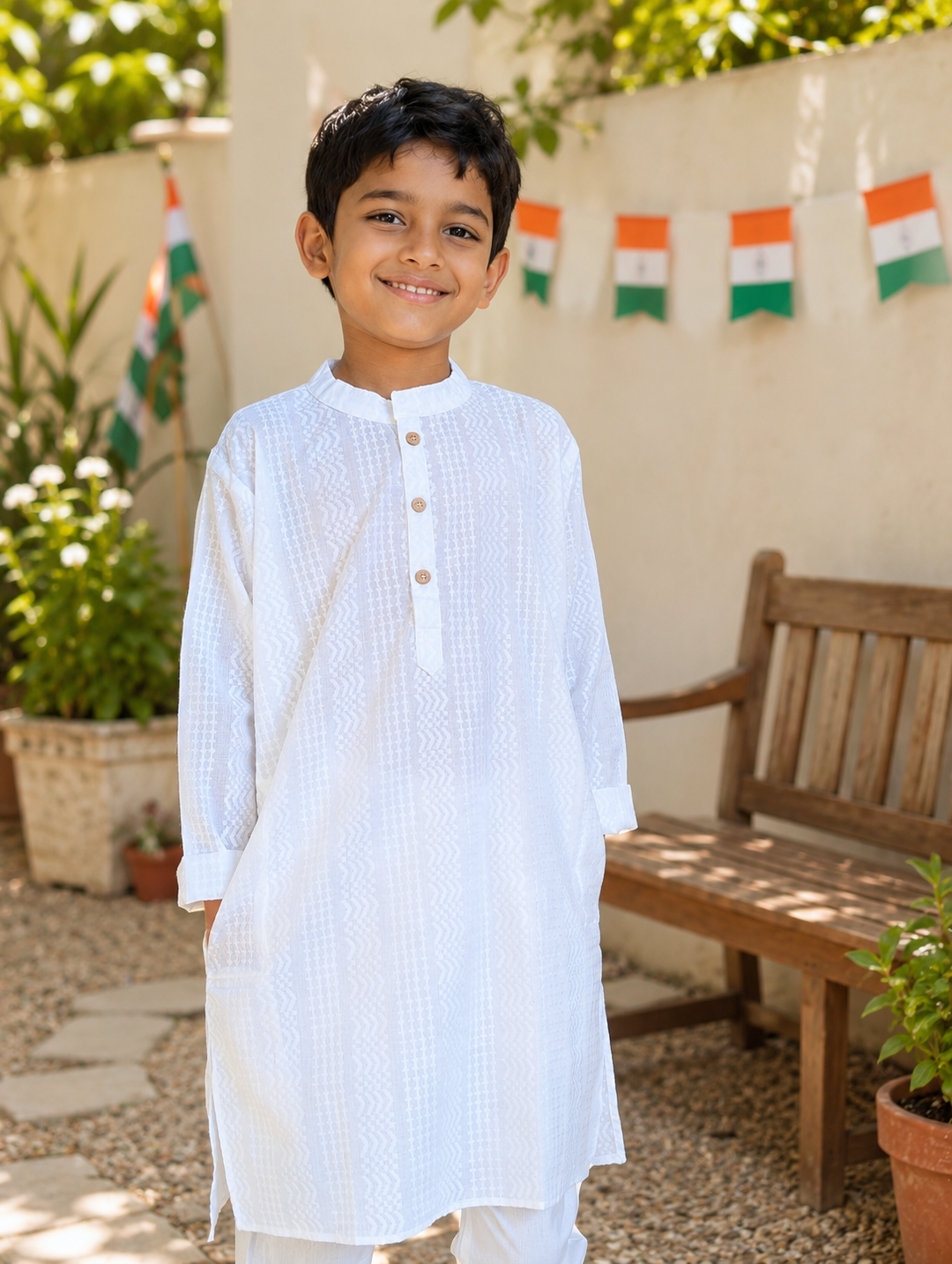 A young boy smiling and wearing a white textured kurta with a three-button placket, standing in an outdoor garden setting with potted plants, a wooden bench, and small Indian flag banners hanging in the background.